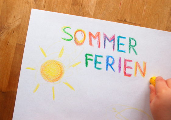 Foto von einem Kind, welches eine Sonne malt und das Wort "Sommerferien" dazu schreibt