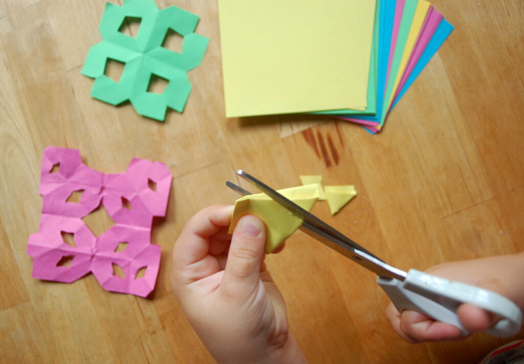Foto von einem Stapel Origami-Papier. Im Vordergrund sind Kinderhände zu sehen, welche mit einer Schere Muster in das Papier schneiden. Symbolbild für Kreativität.